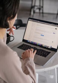 Back view of crop anonymous female remote employee typing on portable computer while watching website on screen and sitting at plastic table with cup of hot drink in apartment